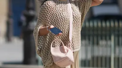 A traveler wears a biege knit fishnet mesh top with wide sleeves, a pastel pink leather shoulder cross body bag, during a street style fashion photo shoot, on August 22, 2026 in Paris, France.