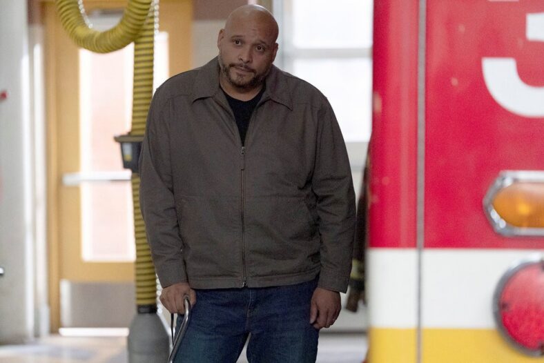 Joe Minoso as Joe Cruz standing inside the firehousewearing a brown jacket with a zipper and jeans while leaning slightly on a stick.