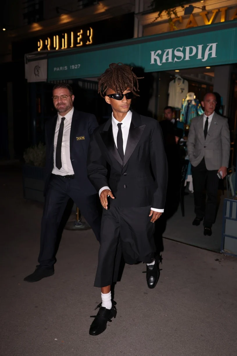 Jaden Smith walks out at night in a cropped black tuxedo jacket, relaxed black trousers, a white shirt, a black tie, white socks, black dress shoes, and slim black sunglasses.